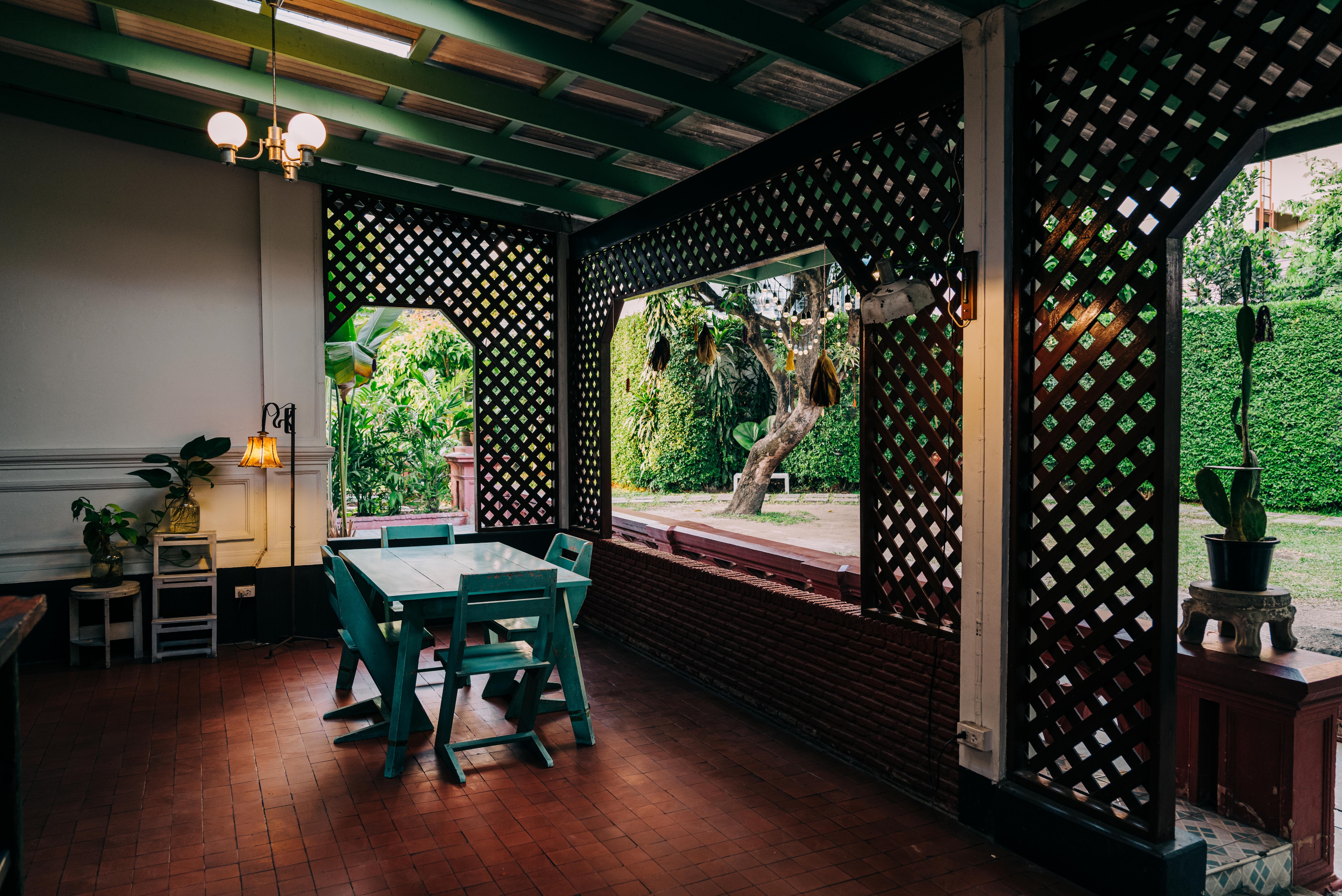 A cafe area shows an aqua green table and chairs with a warm light fixture above and a warm lamp lightning the space, glowing off the warm wooden floor. The space looks like a patio, as it is open to the oudoors with trellis-like walls and large openings. Bright greenery can be seen through the openings.